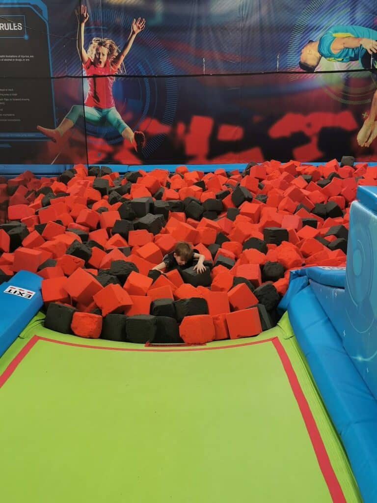 A young child is partially submerged in a large foam pit filled with red and black foam cubes at an indoor trampoline park. The pit is bordered by bright green and blue padded mats. Colourful wall graphics of jumping people are visible in the background.