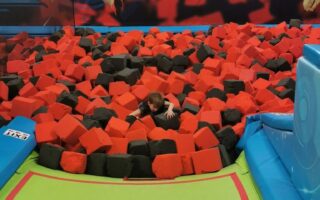 A child is half-buried in a pit filled with red and black foam cubes at an indoor trampoline park. Colourful murals of jumping children are visible on the back wall. Soft green mats and blue padding surround the foam pit for safety.