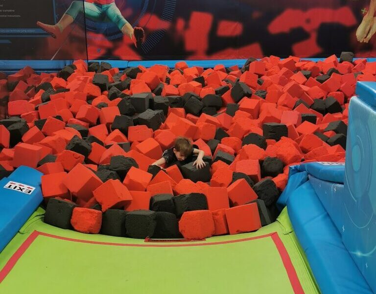 A child is half-buried in a pit filled with red and black foam cubes at an indoor trampoline park. Colourful murals of jumping children are visible on the back wall. Soft green mats and blue padding surround the foam pit for safety.