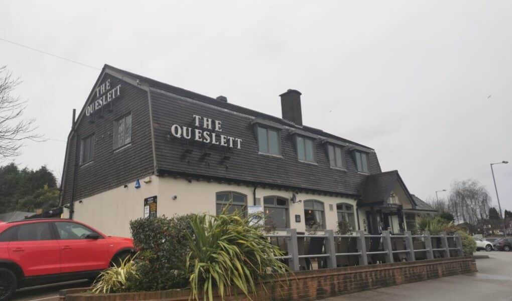 A two-storey building with “THE QUESLETT” sign in large white letters sits on a corner lot. The exterior is tan with dark wood upper walls. A red SUV is parked next to the building, and some shrubs grow along a low brick wall in the foreground.