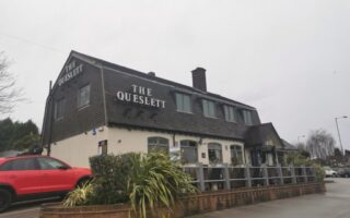 A two-storey building with “THE QUESLETT” sign in large white letters sits on a corner lot. The exterior is tan with dark wood upper walls. A red SUV is parked next to the building, and some shrubs grow along a low brick wall in the foreground.