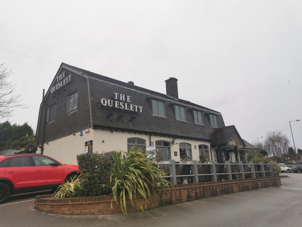 A two-storey pub called “The Queslett” with dark wood panelling on the upper floor and cream-coloured walls below. A bright red SUV is parked out front. There are some plants, outdoor seating, and a cloudy sky above.