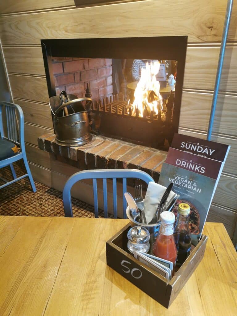 A cosy café scene with a wooden table set for two, holding condiments in a box labelled “50.” Behind the table, there’s a brick fireplace with a blazing fire and a metal kettle, creating a warm, inviting atmosphere. A Sunday menu stands on the table.