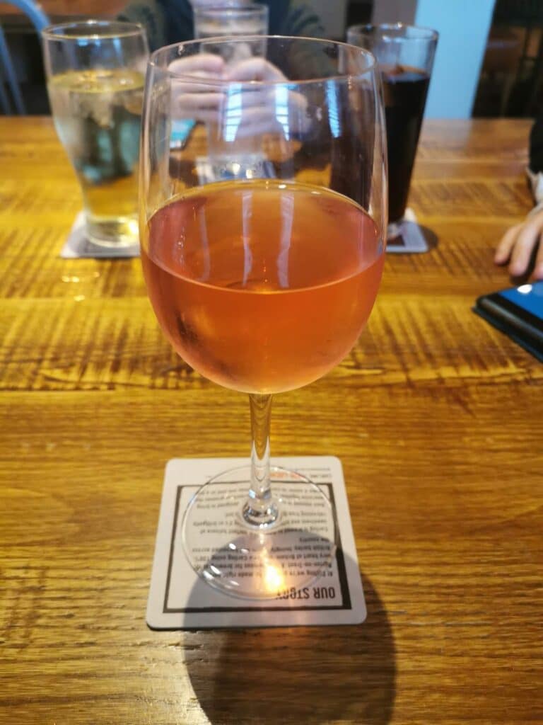 A glass of rosé wine sits on a coaster atop a wooden table. In the background, there are two glasses—one with water and one with a dark beverage. Two hands and a blue mobile are partially visible near the top corners.