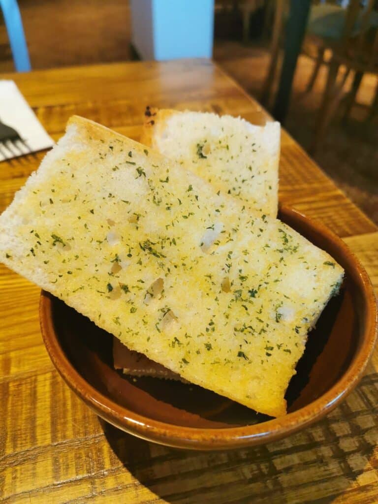 Two rectangular slices of garlic bread topped with herbs are placed in a small brown bowl on a wooden table. In the background, part of a serviette and fork are visible, along with chairs and a blurred interior of a restaurant.