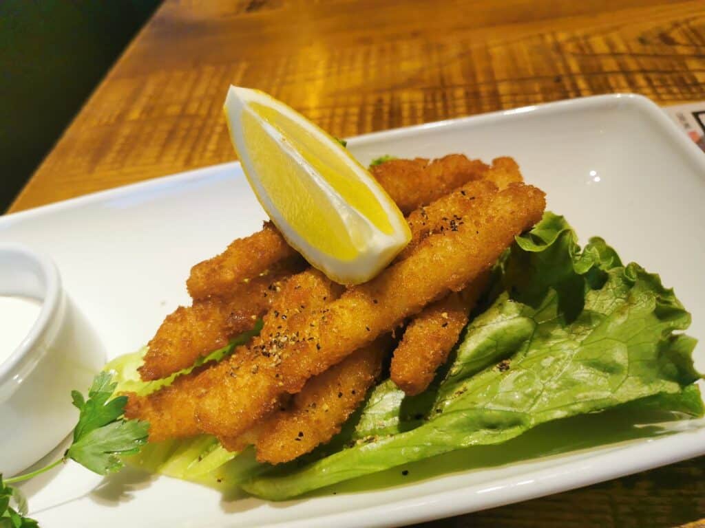 Five golden-brown breaded fish fillets rest on green lettuce leaves on a white plate, topped with a lemon wedge. A small pot of creamy white dipping sauce sits nearby. The plate is set on a wooden table.