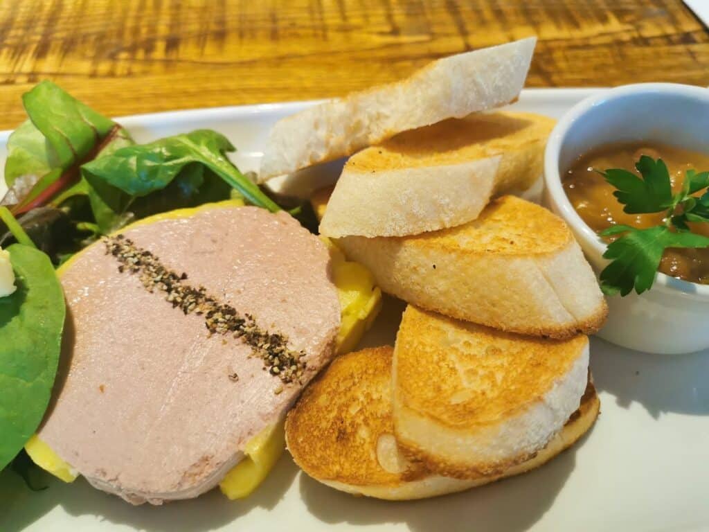 A plate with a slice of liver pâté topped with cracked pepper, toasted baguette slices, a small side of salad leaves, and a ramekin of chutney garnished with parsley, all set on a wooden table.