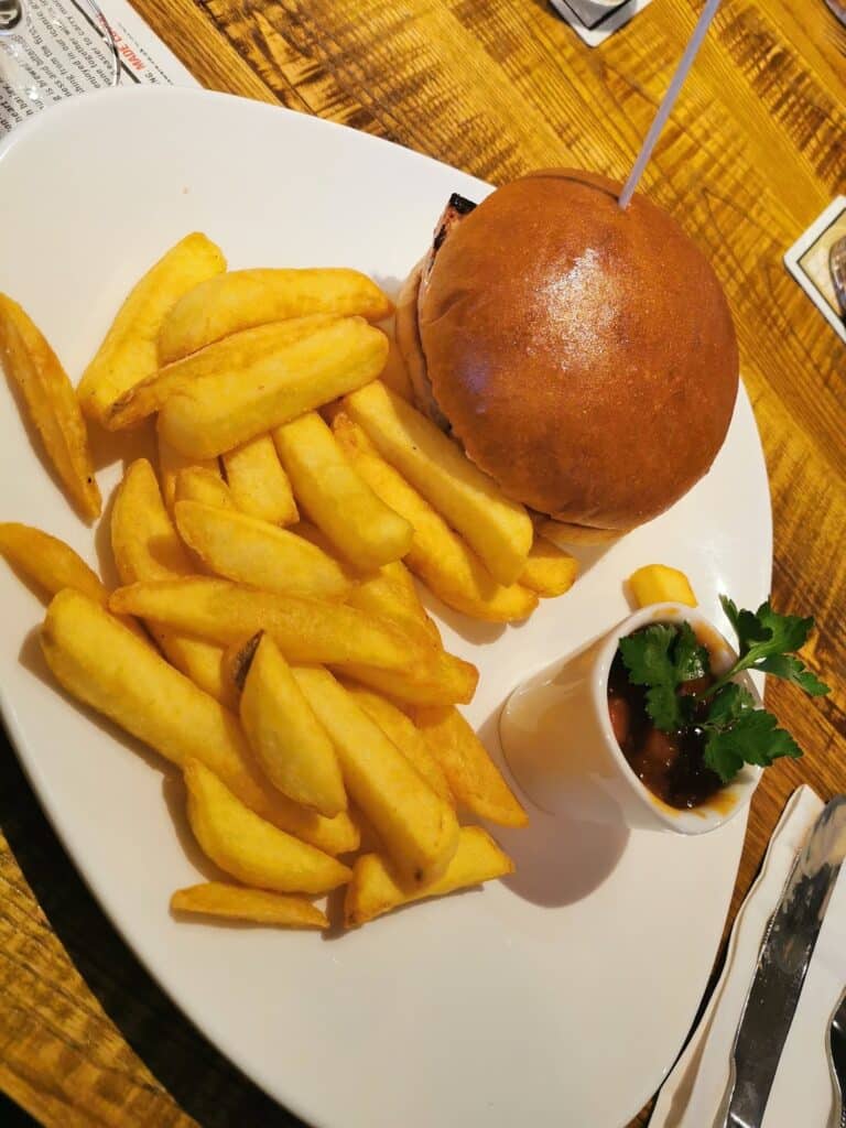 A plate with a burger in a shiny bun, thick golden chips, and a small cup of sauce garnished with parsley. The plate sits on a rustic wooden table with cutlery and part of a menu visible in the background.