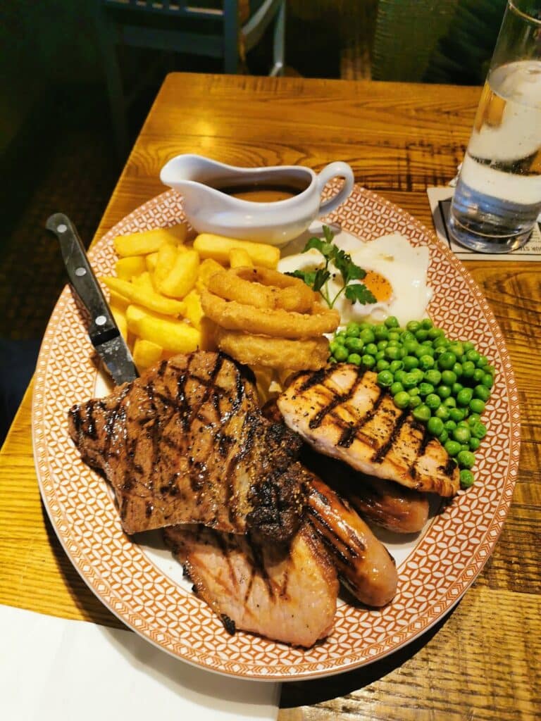 A plate with grilled meats, including chicken and steak, crinkle-cut chips, onion rings, green peas, and a fried egg. A small jug of brown gravy and a glass of water are next to the plate on a wooden table. A knife rests on the plate.