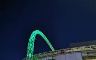 The image shows Wembley Stadium at night, with its signature arch lit up in green against a dark blue sky. The stadium’s exterior is illuminated, with glass panels reflecting the lights, and some streetlights and structures visible in the foreground.