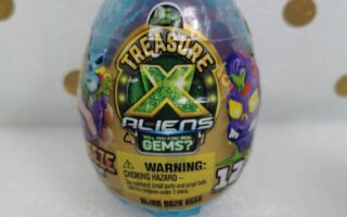 A blue, egg-shaped toy packaging labelled "Treasure X Aliens" sits on a white surface with gold polka dots. Cartoon alien characters and a warning about choking hazards are visible on the front label. The packaging is sealed in clear plastic.