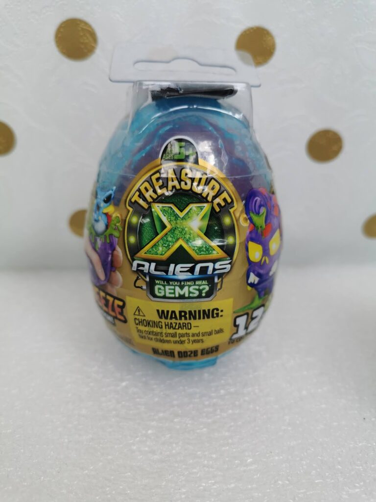 A Treasure X Aliens toy egg with a blue top, purple and green graphics, and gold label. The label features cartoon aliens, the Treasure X logo, and warning text about choking hazards. The background is white with gold polka dots.