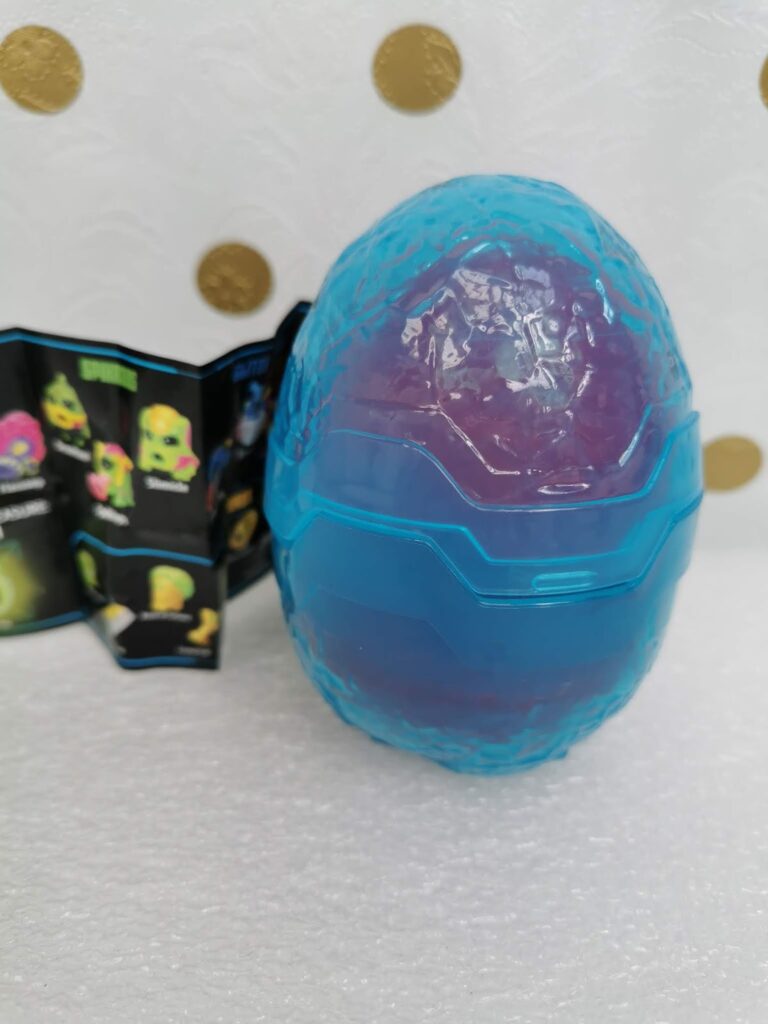 A blue and purple plastic egg-shaped toy with a textured surface sits on a white surface. Behind it, there is a small open leaflet showing colourful cartoon monster faces. The background is white with gold polka dots.