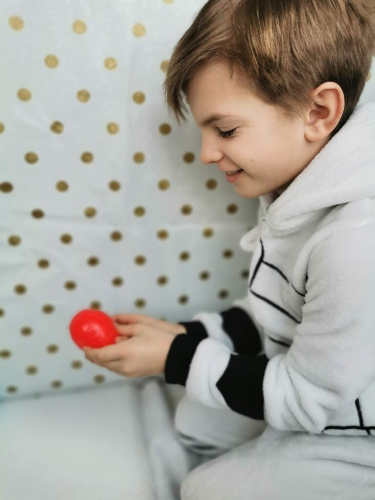 A young child in a white and black striped hoodie sits on a bed, smiling and holding a small red ball. The background features a white blanket with gold polka dots. The child looks focused and happy.