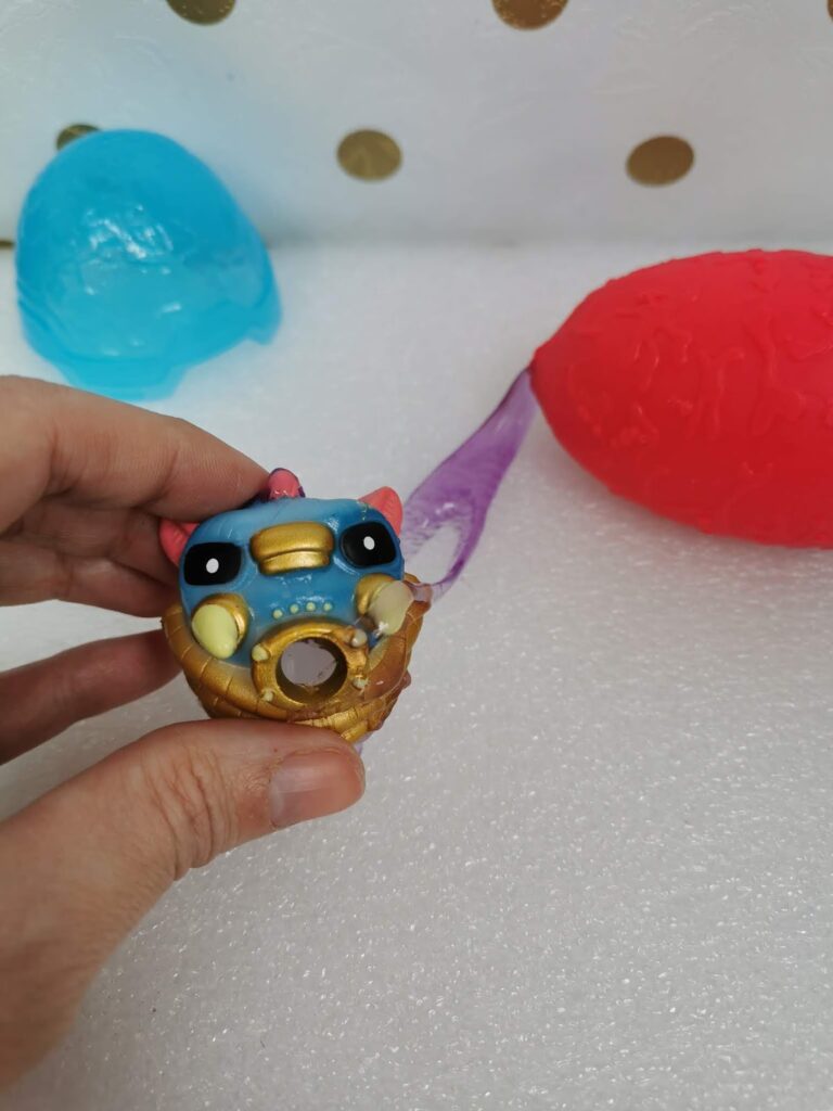 A hand holds a small, round, blue and gold toy with big eyes and a beak, resembling a bird. In the background, there are textured plastic egg toys in blue and red on a white, textured surface with gold polka dots.