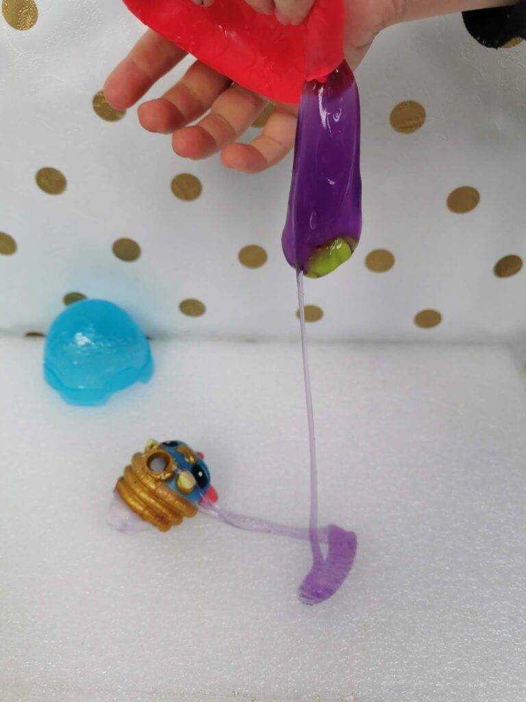 A hand stretches shiny purple slime, which drips over a small toy with a gold and blue top. There is also a blue, dome-shaped toy on a white surface with gold polka dots in the background.