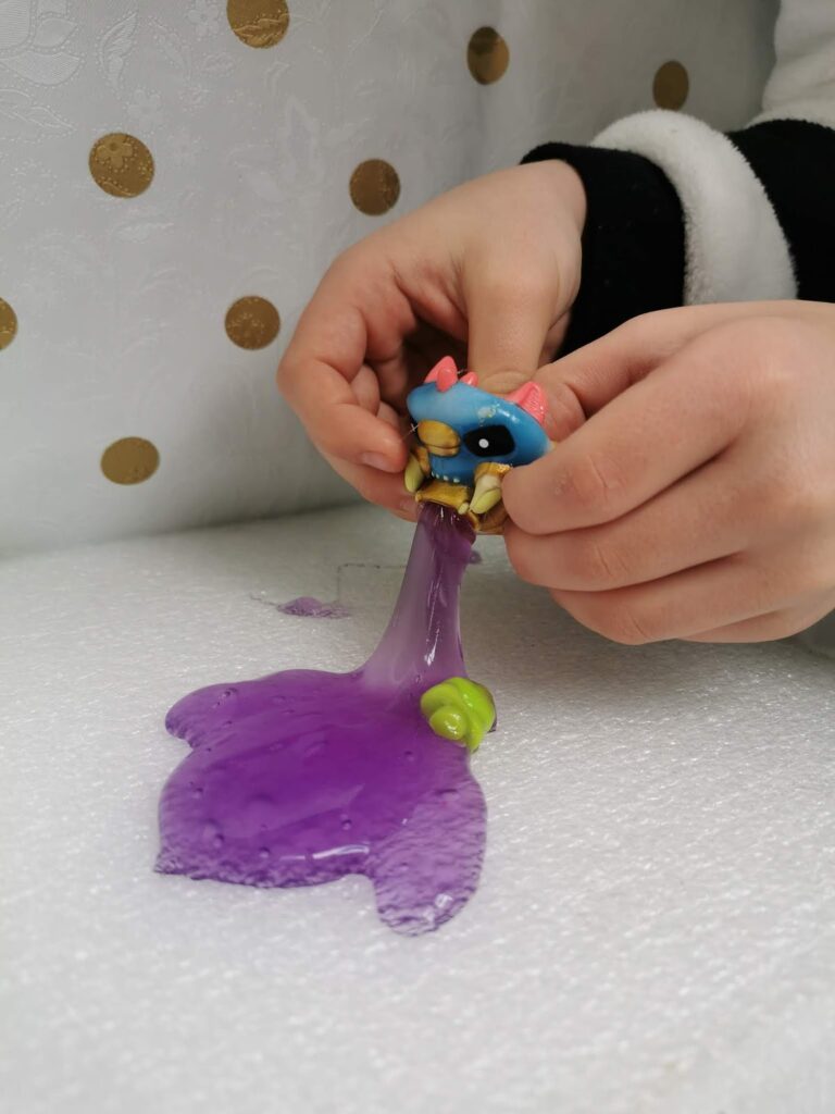 A child’s hands squeeze a small blue monster toy, causing thick purple slime to ooze out of its mouth onto a white textured surface. Gold polka dots are visible on a white background behind the hands. The child is wearing a white sleeve with a black cuff.