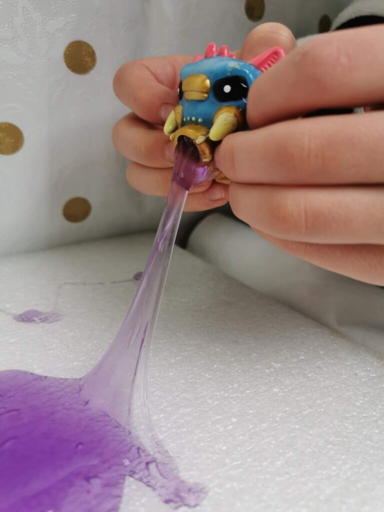 A child squeezes a colourful toy bird, causing purple slime to ooze from its open mouth onto a white surface with gold polka dots in the background. The child’s hands are visible, stretching the slime downward.