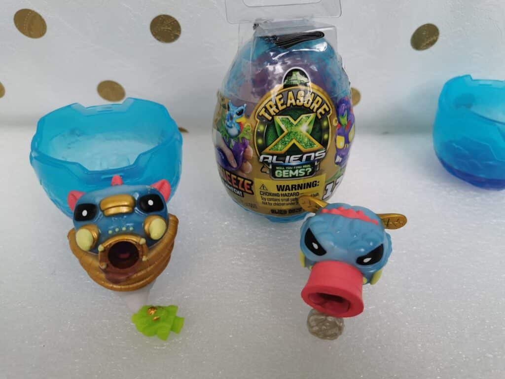 A toy set includes a blue dragon head with an open mouth, a blue and red creature with a pink snout, a small green accessory, a blue plastic egg case, and a packaged Treasure X Aliens Gems egg container on a white surface with gold polka dots.