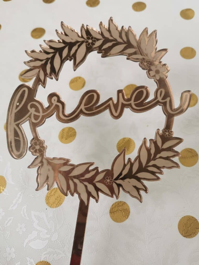 A gold cake topper with the word “forever” in cursive, encircled by leaves and small flowers. It stands upright on a white tablecloth with a gold polka dot pattern.