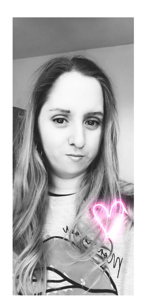Black and white selfie of a woman with long, wavy hair, looking at the camera with a slight smile. She wears a light-coloured top. A pink neon heart graphic is drawn on the lower right side of the image near her shoulder.
