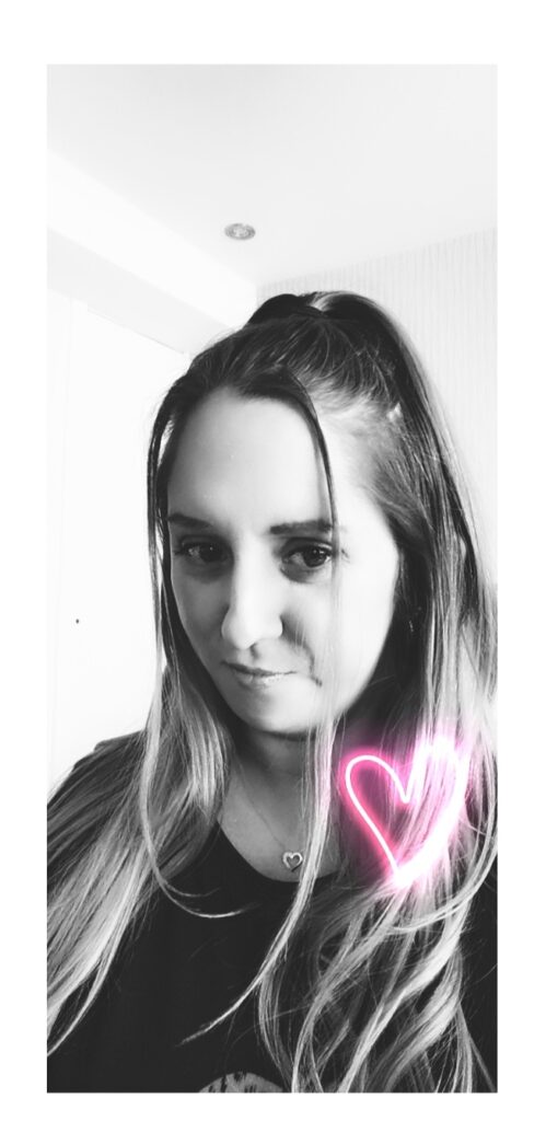 Black and white photo of a young woman with long hair, wearing a dark top and a necklace with a small heart pendant. She is smiling slightly. A pink neon heart graphic is overlaid near her shoulder. The background is a bright indoor setting.