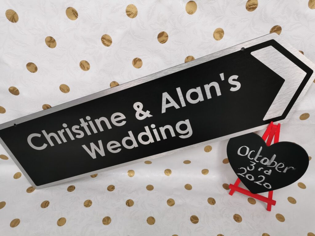 A black sign with white text reads "Christine & Alan's Wedding" and has a white arrow. Next to it is a black heart-shaped board on a red easel, handwritten with "3rd October 2020." The background is white with gold polka dots.