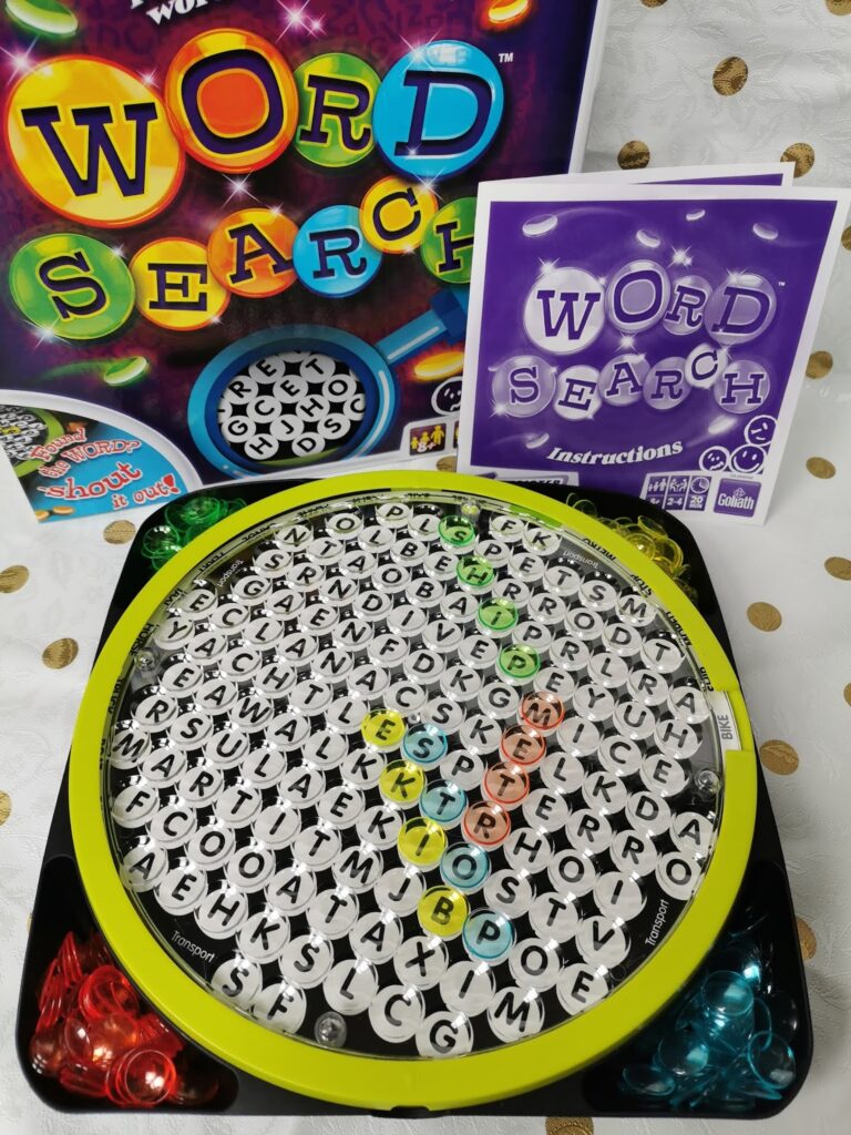 A round Word Search board game with black and white letter tiles, coloured markers highlighting found words, instructions, and the game box in the background. The board and packaging feature a colourful "Word Search" logo and playful design elements.
