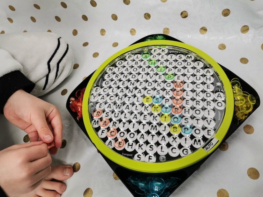 A child’s hands are near a round word search game with multicoloured pegs on each side. The game has a circular grid filled with letters, and some words are highlighted. The surface underneath has a white cloth with gold polka dots.