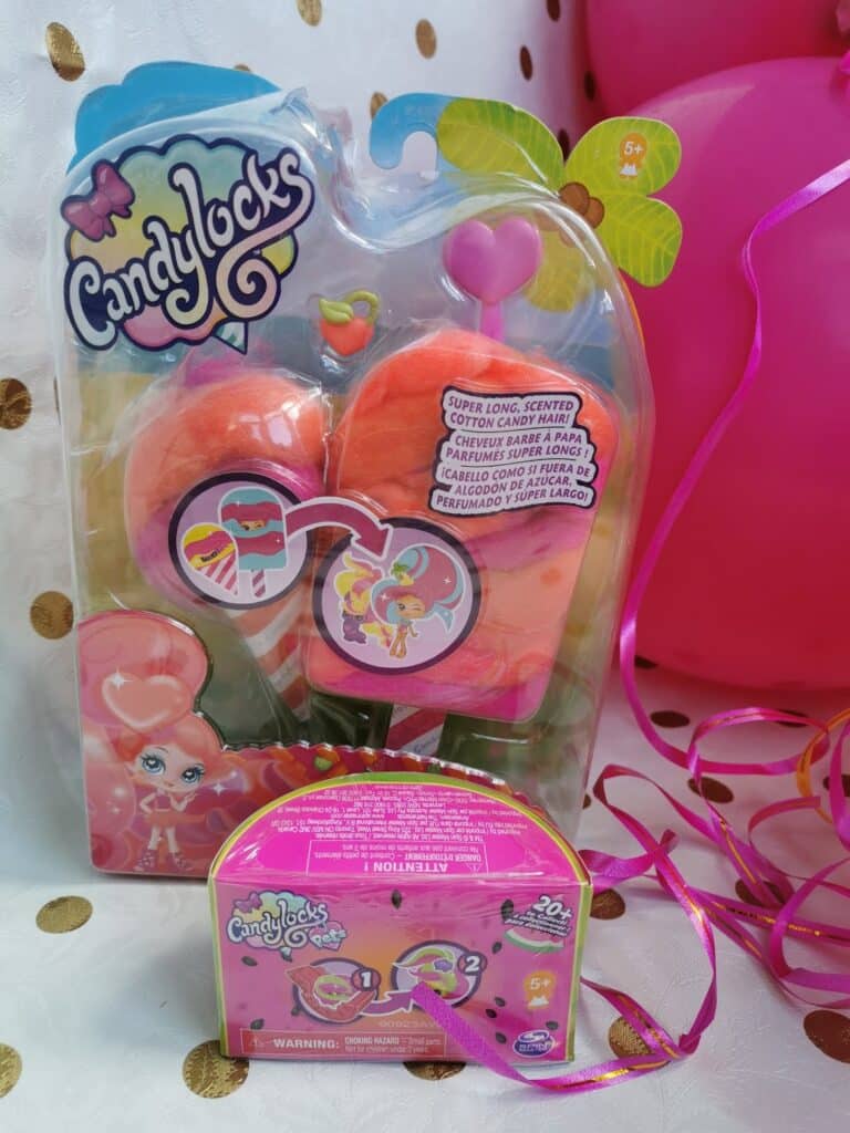 A packaged Candylocks doll with pink and orange hair sits upright against a white and gold polka dot background. Pink balloons and shiny ribbon are on the right. In front, there is a small, closed pink Candylocks accessory box with colourful illustrations.