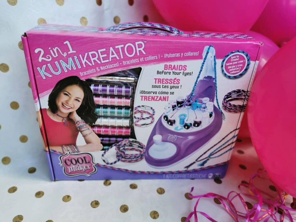 A "2-in-1 KumiKreator" bracelet and necklace making kit by Cool Maker is displayed in its colourful box. The box shows a smiling girl, spools of coloured threads, finished braids, the braiding device, and text in multiple languages. Pink balloons are nearby.