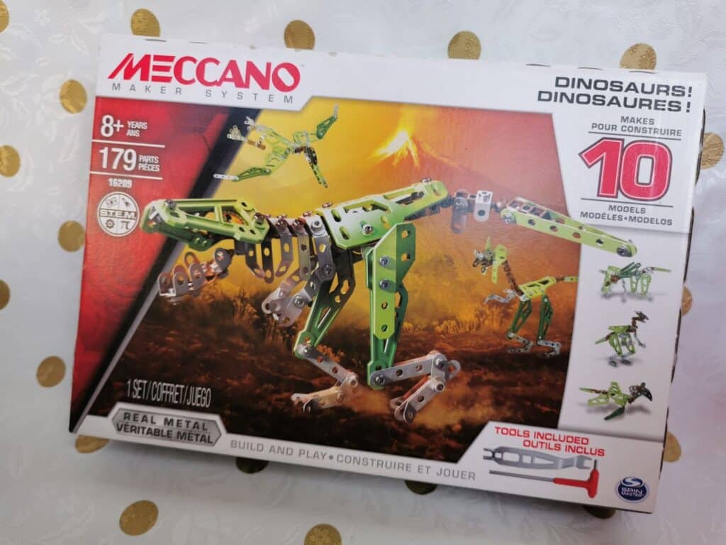 A Meccano Maker System kit box displays a green and silver metal dinosaur model with poseable legs and arms. The box says it makes 10 dinosaur models, contains 179 parts, is for ages 8+, and includes tools. The background is white with gold dots.