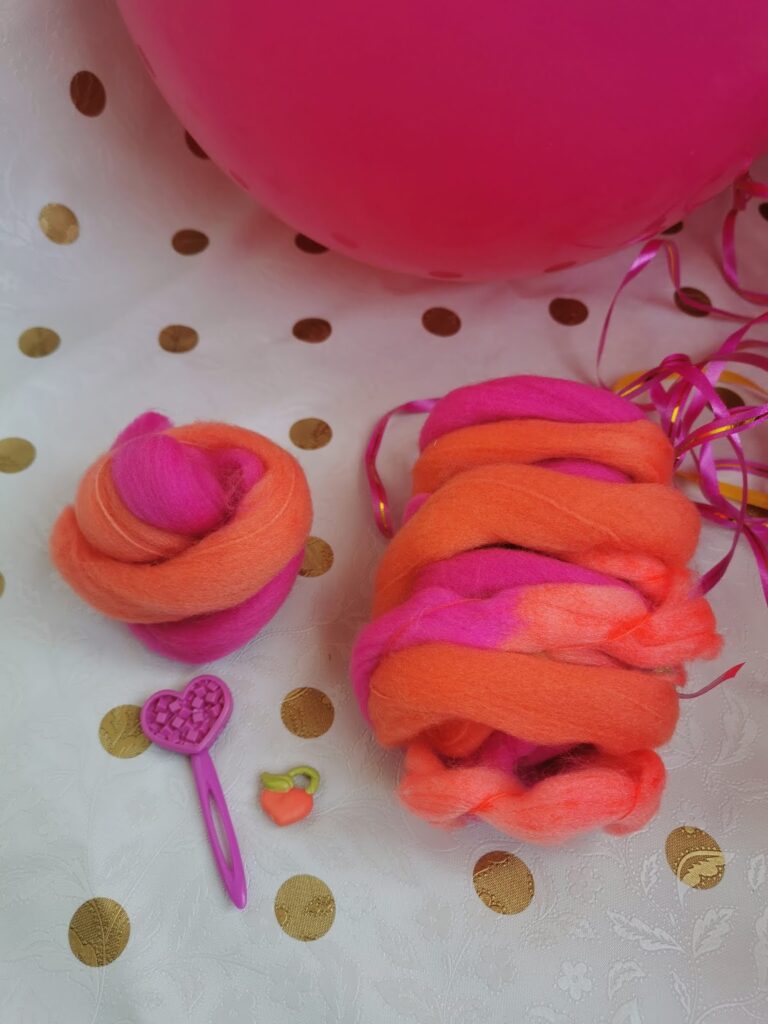 Orange and pink wool roving is displayed on a white tablecloth with gold dots. One bundle is coiled, the other loosely stacked. Nearby are a pink heart-shaped toy brush, a tiny orange charm, and pink curling ribbon beside a large pink balloon.
