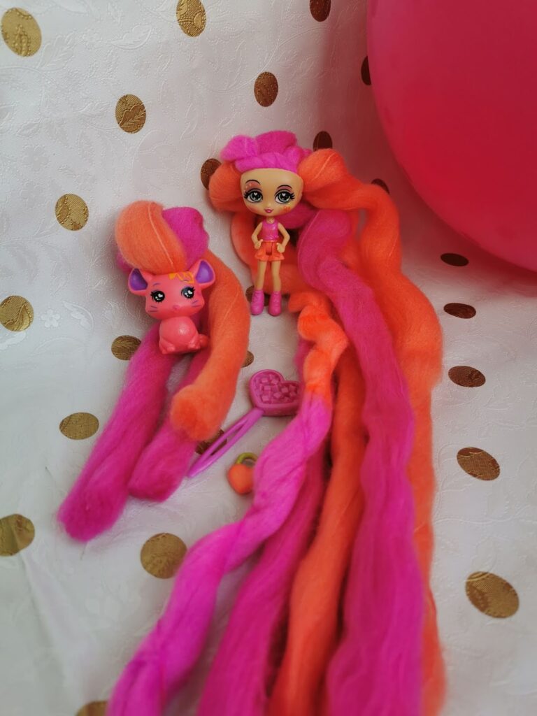 A small doll and a toy animal with long, bright pink and orange hair sit on a white fabric with gold spots. The doll wears a pink outfit and boots. There are toy accessories and a large pink balloon beside them.