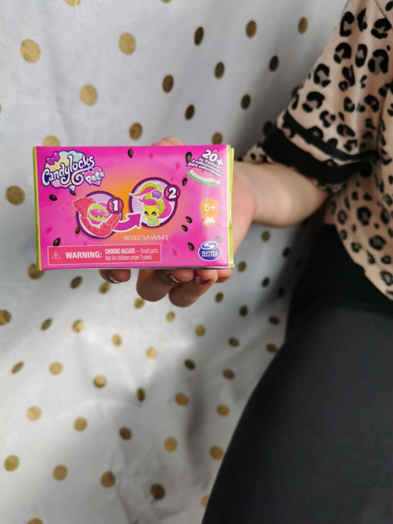 A person holds a colourful Candylocks toy box with cartoon illustrations of the toy’s contents. The box is pink and green with images of two collectable figures. Gold polka dot fabric is in the background, and the person wears black trousers and a leopard print shirt.