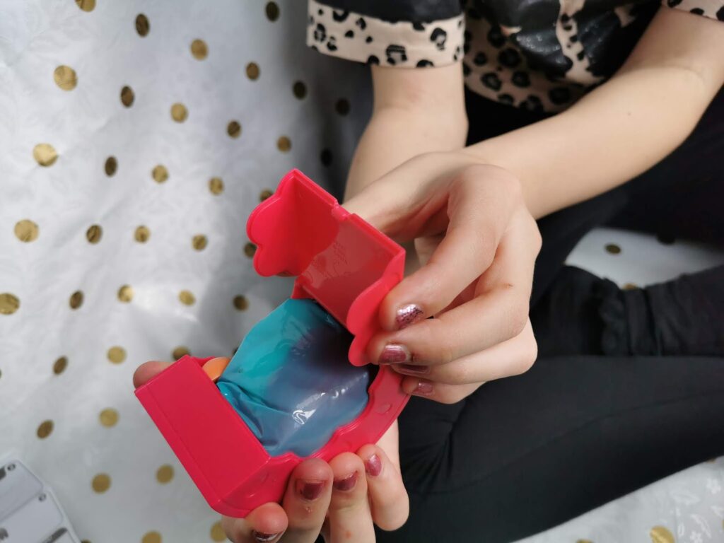 A child with painted fingernails holds an open red plastic toy container with blue slime inside. The child is sitting on a white surface with gold polka dots, wearing black trousers and a white shirt with a black pattern.