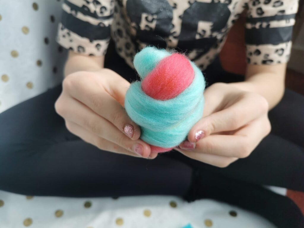 A person with glittery nail varnish holds a swirl of soft wool in blue and pink. They wear black leggings and a beige shirt with a bold black print. The background includes a white and gold polka-dotted surface.