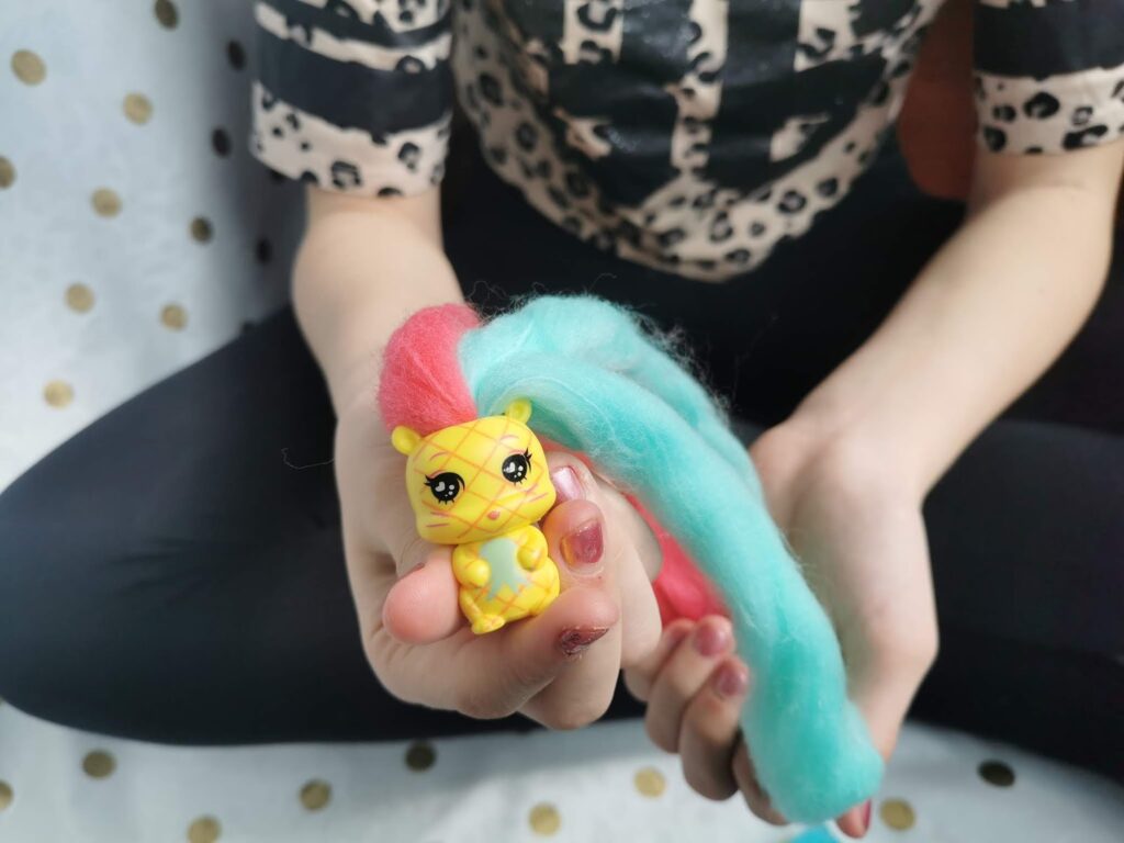 A child with fair skin, wearing a black and beige animal print shirt and black trousers, sits cross-legged holding a small yellow toy animal with a red and blue mane of fuzzy hair in their right hand. The background is white with gold polka dots.