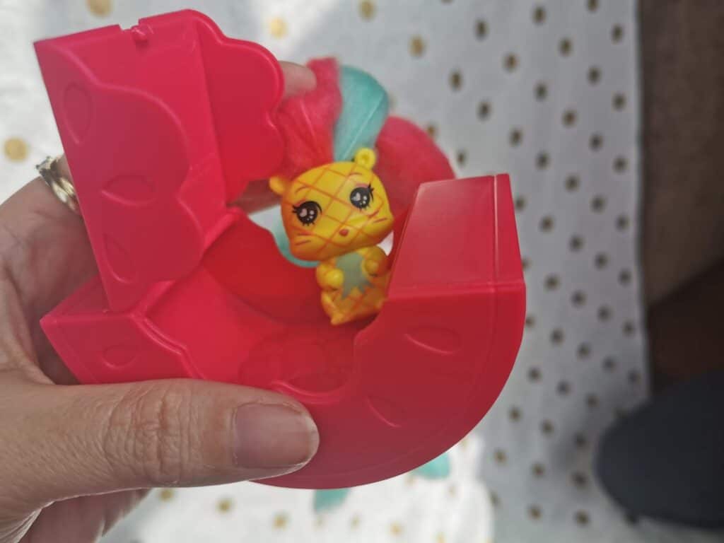 A hand holds a red, semi-circular plastic toy case that is open, revealing a small yellow toy animal with orange stripes, large eyes, and a teal-and-pink mane inside. The background is a white fabric with gold polka dots.