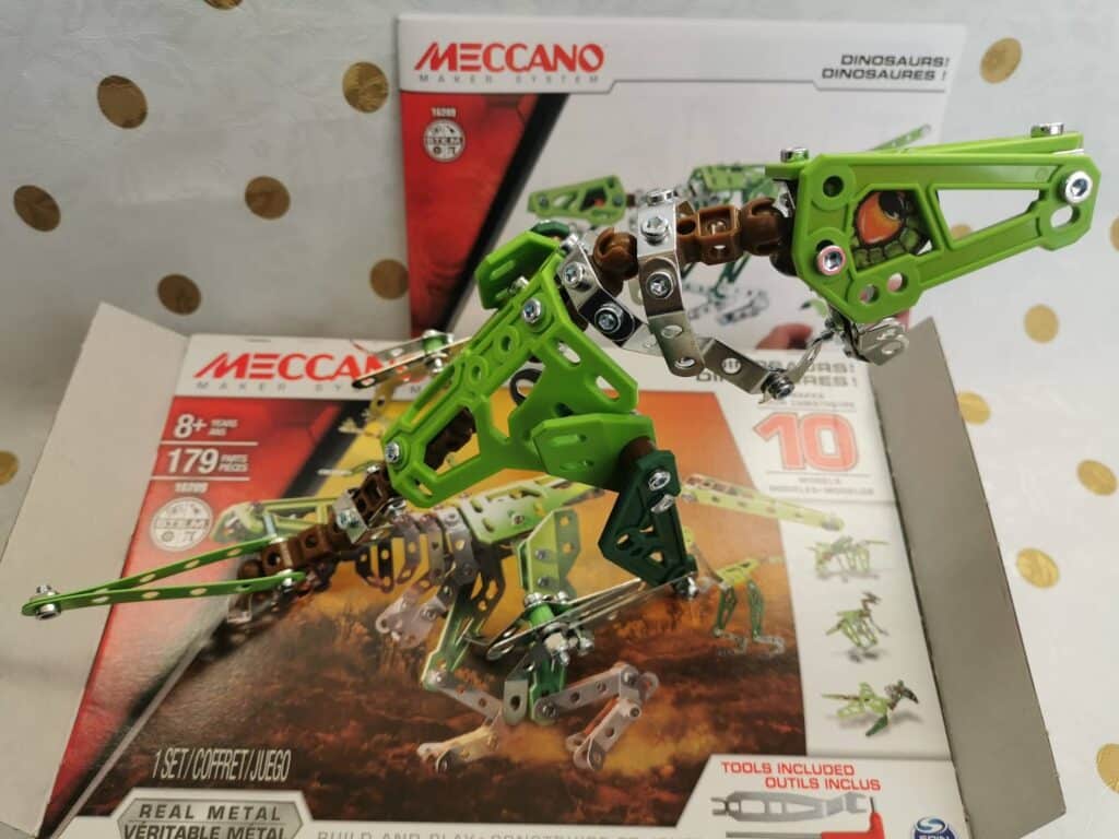 A green and silver metal dinosaur model, assembled from Meccano parts, stands on its box. The packaging displays the Meccano logo, an image of the dinosaur, and text indicating 179 pieces, real metal, for ages 8+, and that tools are included.