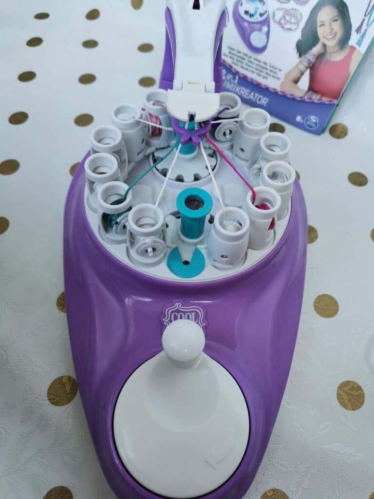 A purple bracelet-making machine is placed on a white table with gold dots. Colourful threads are looped through the device. In the background, the toy’s packaging shows a smiling girl and a sample bracelet, suggesting it is used for crafting jewellery.