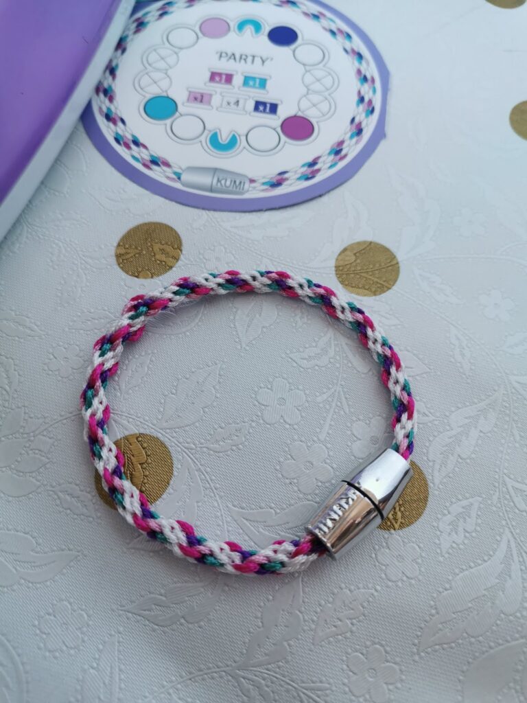 A plaited friendship bracelet with pink, blue, black, and white threads lies in a circle on a textured white surface with gold dots. The bracelet has a metallic clasp. Behind it is a round Kumi bracelet pattern card with coloured guides and the word "PARTY.