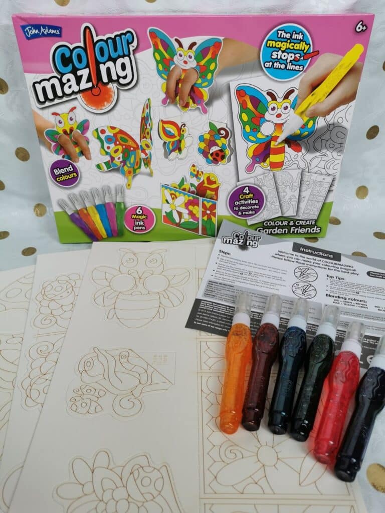 A Colourmazing Garden Friends art kit is displayed with the box, four blank illustrated sheets, a sheet of instructions, and six coloured ink pens—yellow, orange, red, blue, green, and black—arranged on a white surface with a gold polka-dot background.