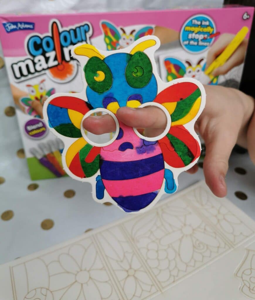 A child’s hand holds a brightly coloured paper bee mask with finger holes, coloured in rainbow hues. In the background is a Colour Mazing art kit box showing a similar bee design. The tablecloth below is white with gold dots.