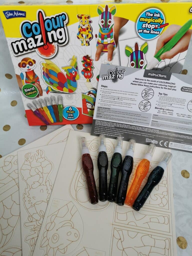 A Colourmazing art kit is shown with its box, six coloured pens, three sheets of animal-themed colouring pages with outlines, and an instruction leaflet. The box features colourful finished animal designs and promotes "magic ink" that stops at the lines.
