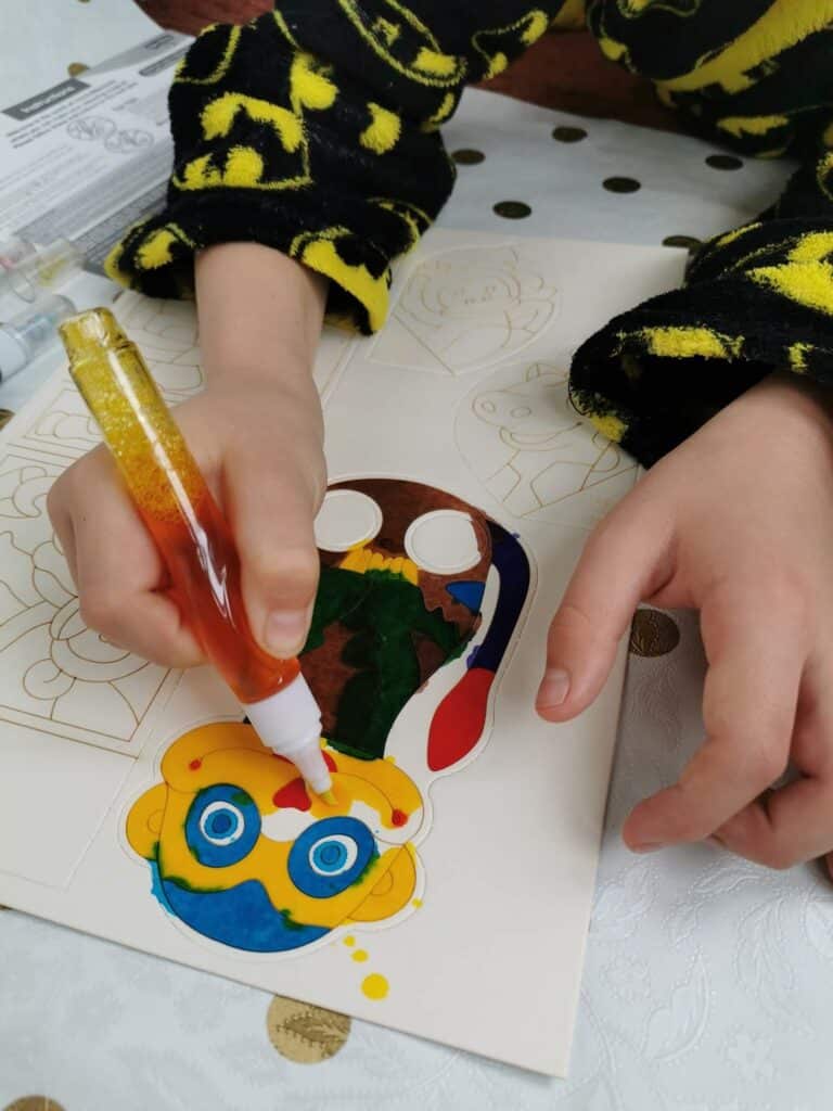 A child wearing a black and yellow Batman-patterned dressing gown uses a paint pen to colour a cartoon figure with blue glasses, yellow face, and multicoloured outfit on a colouring sheet resting on a white table with gold dots.