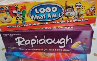 Three colourful board game boxes are stacked. The top box is "LOGO What Am I?"; the middle is "Rapidough" showing playdough and a hand; the bottom is "Articulate! For Kids." The background is white with gold polka dots.