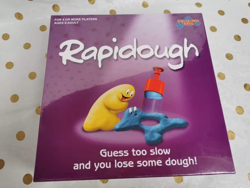 A purple game box titled "Rapidough" features colourful dough characters—a yellow one with a face and a blue one shaped like a star. Text reads, "Guess too slow and you lose some dough!" The box is on a white table with gold polka dots.