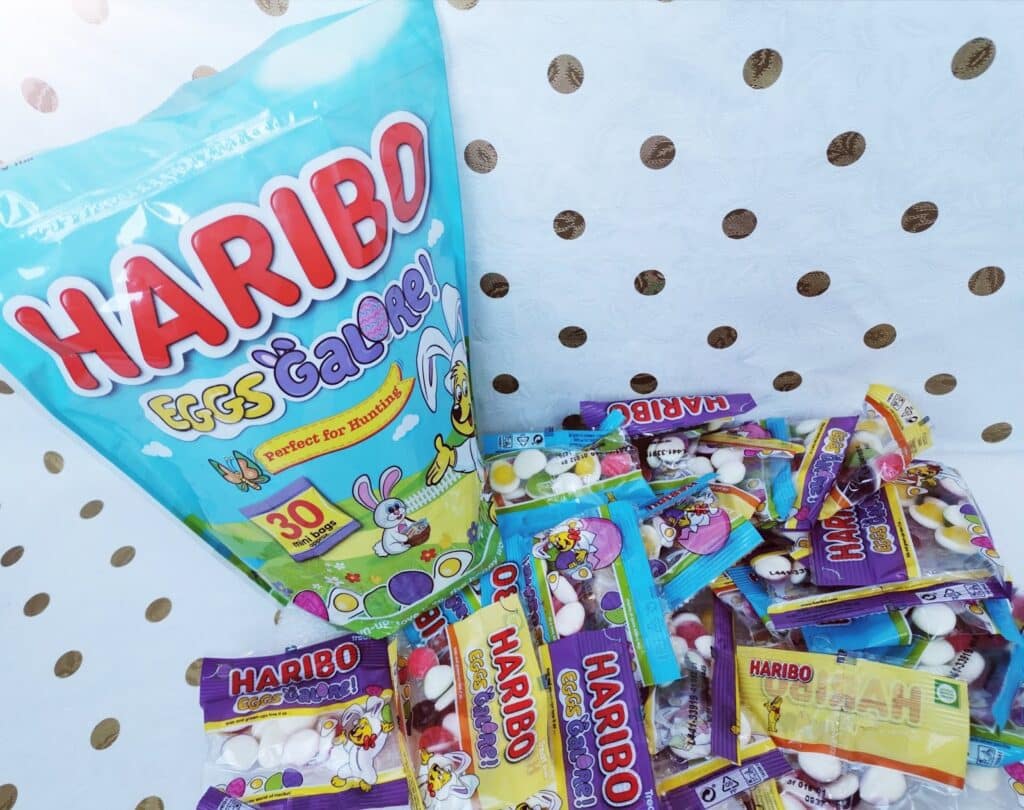 A large blue bag labelled "Haribo Eggs Galore!" with a cartoon bunny sits on white paper with gold dots. Around it are several small packs of Haribo gummy sweets, each filled with colourful gummies shaped like eggs.