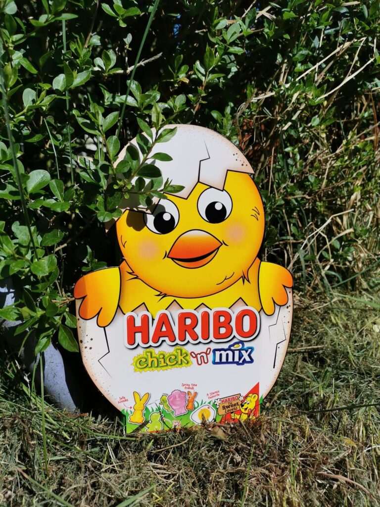 A colourful metal tin shaped like a yellow chick hatching from an egg, labelled "HARIBO Chick ‘n’ Mix," sits on grass among green bushes. Cartoon sweets are illustrated at the bottom of the tin. Sunlight highlights the scene.
